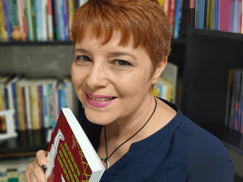 Professora Denise Ferreira sorrindo, tendo uma estante com livros ao fundo. Ela segura um livro sobre linguagem descritiva.